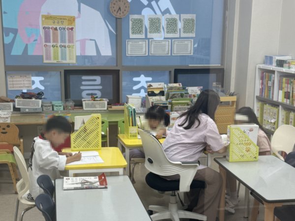 공부하는코끼리수학 초등 1~2학년 교실. 아이들이 각자 다른 교재로 문제를 풀고, 선생님이 옆에 앉아 한 아이를 봐주고 있다. 뒤쪽 게시판에 구구단표와 학습 자료가 붙어 있다. 학생 얼굴은 개인정보 보호를 위해 가렸다.